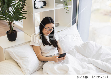 Asian woman relaxing in bed using smartphone, possibly texting or browsing social media, in bright morning light. Comfortable home environment with casual white clothing. Asian woman relaxing in bed using smartphone, possibly texting or browsing social media, in bright morning light. Comfortable home environment with casual white clothing. 121957571