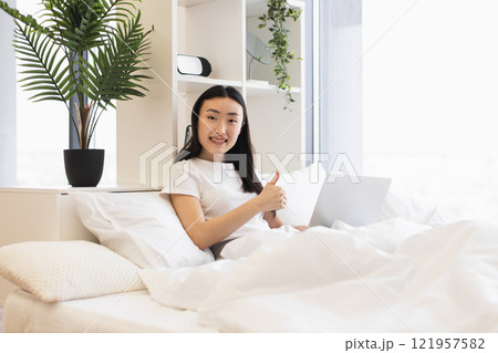 Asian woman lounging in bed with laptop, showing thumbs up. Young adult in white room enjoying comfort and technology. Bright setting with plants and modern decor. Asian woman lounging in bed with laptop, showing thumbs up. Young adult in white room enjoying comfort and technology. Bright setting with plants and modern decor. 121957582