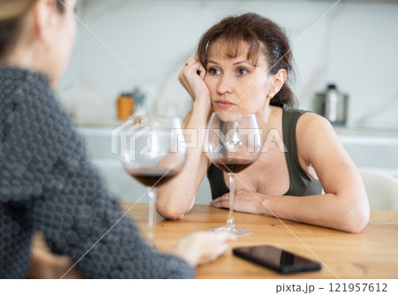 Middle-aged woman sitting at table with a glass of wine and talking to another at home 121957612