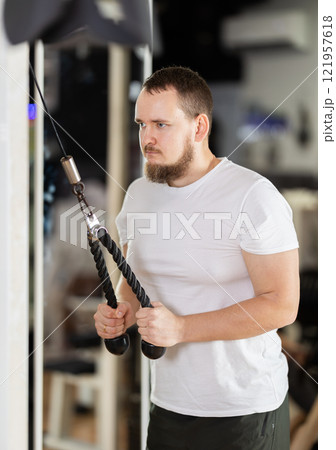 Man in gym performs exercises for arm muscles using cable crossover machine and pull weighted rope 121957618