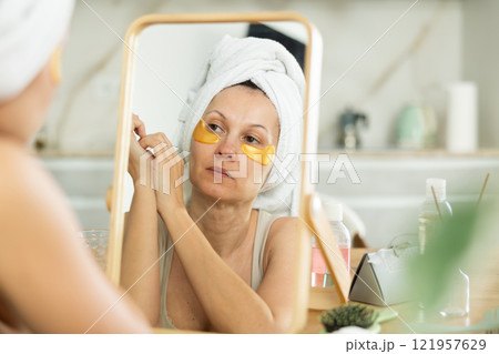 Middle-aged woman applying eye patch mask on her face sitting in front of the mirror Middle-aged woman applying eye patch mask on her face sitting in front of the mirror 121957629