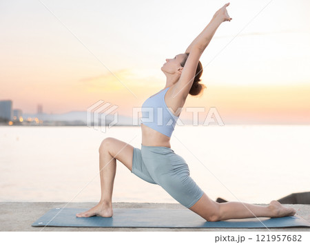 Sporty energetic female teacher doing exercises on pilates mat and demonstrating variation of warrior pose of yoga on beach at sunset. Fitness concept 121957682