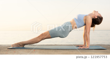 Sporty energetic experienced young woman yoga trainer performing upward plank pose on rug on beach at sunrise. Fitness concept Sporty energetic experienced young woman yoga trainer performing upward plank pose on rug on beach at sunrise. Fitness concept 121957753