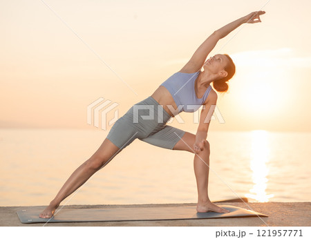 Sporty woman practicing yoga outside, doing relaxing stretching workout for body flexibility on seashore at sunrise 121957771