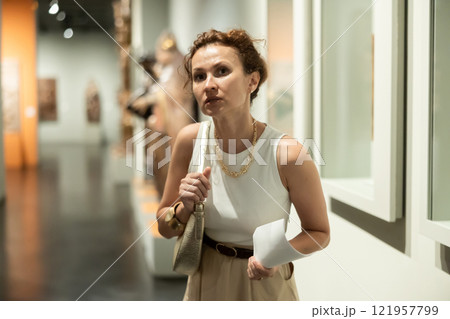 Portrait of middle-aged woman with brochure in her hands examining antique objects in historical museum 121957799
