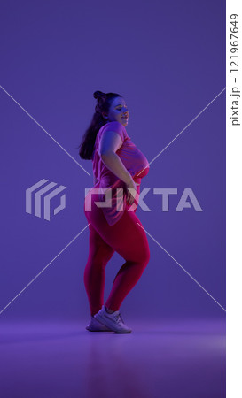 Side view photo of young plus size woman dressed red sportswear posing confidently against purple studio background in vibrant neon light. 121967649