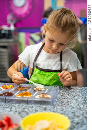 Little girl learning how to cook in a cooking class. Handmade dessert. Making chocolate candy, Child filling heart shaped mold with chocolate 121968482