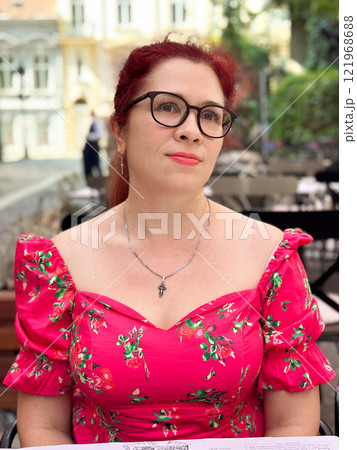 Portrait confident woman in a bright pink floral dress sits outdoors, looking with a calm yet poised expression. Millennial female and summer lifestyle Portrait confident woman in a bright pink floral dress sits outdoors, looking with a calm yet poised expression. Millennial female and summer lifestyle 121968688