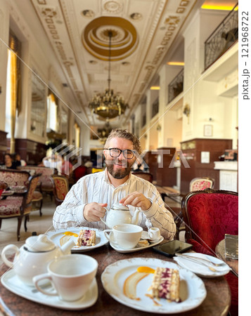 Millennial man enjoying an elegant tea break in a luxurious cafe with classical decor. Luxury restaurant paired with fine tableware and a slice of cake, timeless and refined atmosphere 121968722