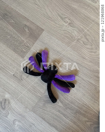 Plush spider toy with purple and black legs rests on a light wooden floor, creating a whimsical and playful atmosphere. Fun and creativity in everyday life Plush spider toy with purple and black legs rests on a light wooden floor, creating a whimsical and playful atmosphere. Fun and creativity in everyday life 121968868