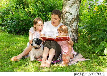 Mother reading book to daughters outdoors with pet dog 121969937