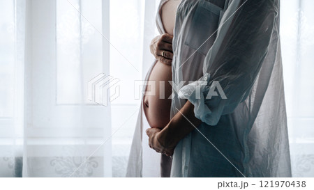 Happy pregnant woman tenderly holding her belly while standing near transparent curtain. Family, pregnancy, child health and motherhood Happy pregnant woman tenderly holding her belly while standing near transparent curtain. Family, pregnancy, child health and motherhood 121970438
