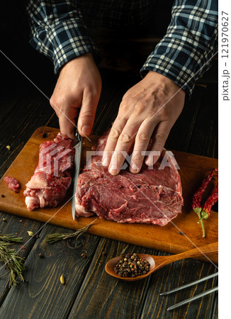 A pair of hands slice through a large piece of fresh meat on a wooden cutting board. Surrounding ingredients like herbs and spices create a warm, inviting atmosphere typical of a cozy kitchen A pair of hands slice through a large piece of fresh meat on a wooden cutting board. Surrounding ingredients like herbs and spices create a warm, inviting atmosphere typical of a cozy kitchen 121970627