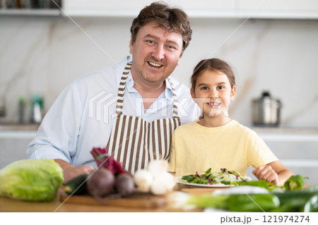 Portrait of father with his little daughter in the kitchen - daughter helps father prepare vegetable salad Portrait of father with his little daughter in the kitchen - daughter helps father prepare vegetable salad 121974274