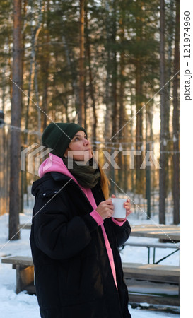 Relaxed young woman in warm winter clothes drinking hot tea outdoors in the backyard of her country house. Nice woman holding a mug with a drink, dreaming, admiring the winter landscape, time for Relaxed young woman in warm winter clothes drinking hot tea outdoors in the backyard of her country house. Nice woman holding a mug with a drink, dreaming, admiring the winter landscape, time for 121974960