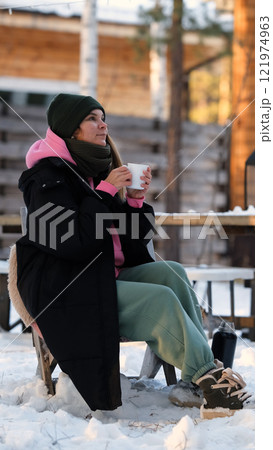 Relaxed young woman in warm winter clothes drinking hot tea outdoors in the backyard of her country house. Nice woman holding a mug with a drink, dreaming, admiring the winter landscape, time for Relaxed young woman in warm winter clothes drinking hot tea outdoors in the backyard of her country house. Nice woman holding a mug with a drink, dreaming, admiring the winter landscape, time for 121974963