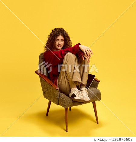 Confident, attractive woman, dressed casual clothes, sitting on chair and looking at camera against yellow studio background. 121976608