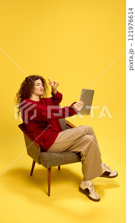 Cheerful smiling young woman sitting in chair and taking selfie on tablet gesturing peace sign against yellow studio background. Cheerful smiling young woman sitting in chair and taking selfie on tablet gesturing peace sign against yellow studio background. 121976614