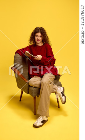 Confident woman, with curly hair, wearing casual clothes, sitting in chair with tablet and looking at camera against yellow studio background. Confident woman, with curly hair, wearing casual clothes, sitting in chair with tablet and looking at camera against yellow studio background. 121976617