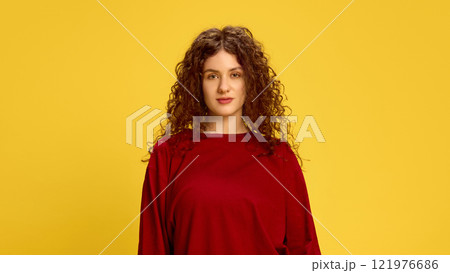 Calm and serious, young woman, with curly brunette hair, dressed red t-shirt, looking at camera against vibrant yellow background. Headshot. Calm and serious, young woman, with curly brunette hair, dressed red t-shirt, looking at camera against vibrant yellow background. Headshot. 121976686