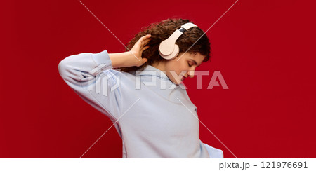 Young, creative girl, with curly brunette hair listening music in headphones against red studio background. Carefree. Young, creative girl, with curly brunette hair listening music in headphones against red studio background. Carefree. 121976691