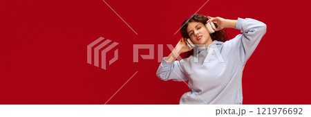 Banner, Young beautiful woman dressed casually holding hands on headphones while listening music against red background with negative space. Banner, Young beautiful woman dressed casually holding hands on headphones while listening music against red background with negative space. 121976692