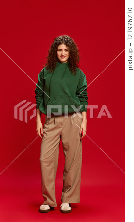Full-body photo of young curly woman, dressed casually, looking with slight smile at camera against bold red studio background. 121976710