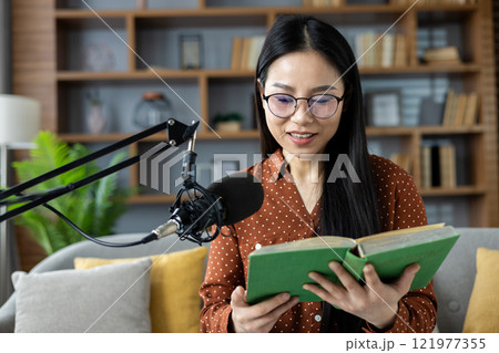 Asian woman with glasses recording podcast at home, reading book. Engaged in creative broadcasting. Relaxed indoor setting, communicating ideas, sharing knowledge. Asian woman with glasses recording podcast at home, reading book. Engaged in creative broadcasting. Relaxed indoor setting, communicating ideas, sharing knowledge. 121977355