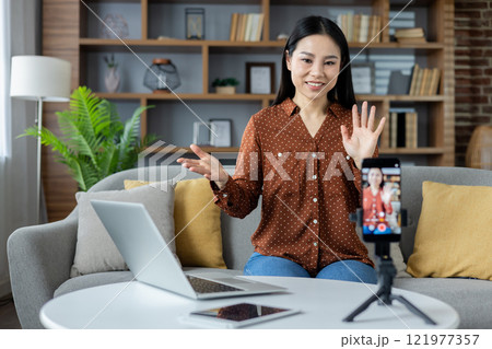 Asian woman engaging in video conference at home. Sitting on sofa with laptop and phone tripod. Modern living room setting. Concept of remote work, online communication. 121977357