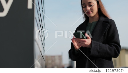 Woman receiving parcel from post terminal machine using smartphone outdoors. Parcel delivery machine. Mail delivery and post service, online shopping, e commerce concept. Woman receiving parcel from post terminal machine using smartphone outdoors. Parcel delivery machine. Mail delivery and post service, online shopping, e commerce concept. 121978074