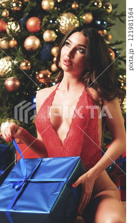 Sexy brunette in red lace bodysuit posing near Christmas tree. Gift satin bow tied around woman's waist. Fashion portrait of model girl in lace lingerie indoors with Christmas tree. Woman posing for Sexy brunette in red lace bodysuit posing near Christmas tree. Gift satin bow tied around woman's waist. Fashion portrait of model girl in lace lingerie indoors with Christmas tree. Woman posing for 121981564