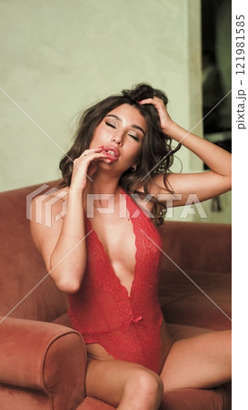 Beautiful brunette posing sexy on camera in red lace bodysuit while sitting on sofa Beautiful brunette posing sexy on camera in red lace bodysuit while sitting on sofa 121981585