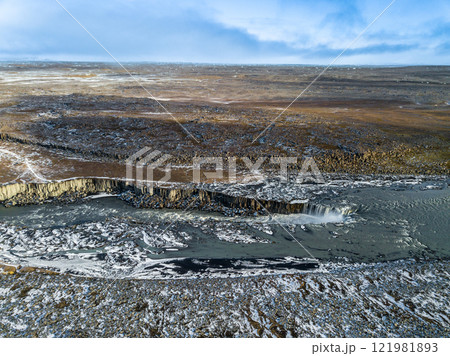 Turquoise glacial river winds through a frozen Icelandic landscape, flowing toward the majestic Dettifoss waterfall, showcasing nature's raw beauty and power from above 121981893