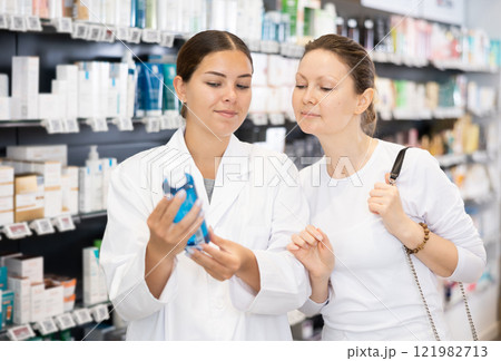 Female pharmacist advises a woman visitor a good skin care remedy, standing in the sales hall of pharmacy Female pharmacist advises a woman visitor a good skin care remedy, standing in the sales hall of pharmacy 121982713