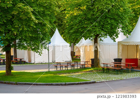 A Row of Elegant White Party Tents Set Up in a Beautiful Park Surrounded by Lush Green Trees. 121983393