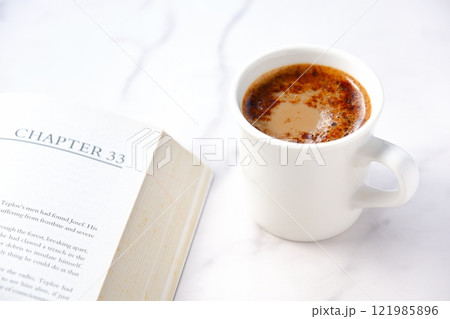 ページを開いたペーパーバック(洋書)とコーヒーの入ったコーヒーカップで、コーヒーを飲みながら読書中 ページを開いたペーパーバック(洋書)とコーヒーの入ったコーヒーカップで、コーヒーを飲みながら読書中 121985896