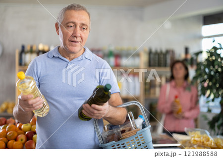 Mature man buyer looks thoughtfully at bottles of vegetable oil, woman client in background Mature man buyer looks thoughtfully at bottles of vegetable oil, woman client in background 121988588
