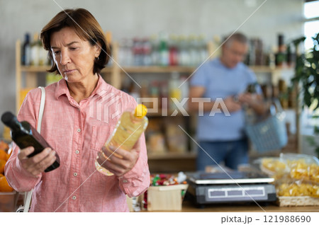 Mature woman buyer looks thoughtfully at bottles of vegetable oil, man client in background Mature woman buyer looks thoughtfully at bottles of vegetable oil, man client in background 121988690