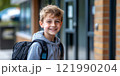 A cheerful young boy smiles brightly while standing outside a school building. His backpack is ready for a day of learning and fun. This image captures the joy of youth and education. AI 121990204