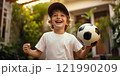 A cheerful young boy wearing a cap joyfully holds a soccer ball in his hands. The bright background features lush greenery, perfect for an outdoor sports theme. A simple and natural style embodies 121990209