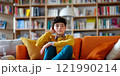 A thoughtful boy sits on a vibrant orange couch in a cozy living room. Shelves filled with books serve as a backdrop. This style captures the essence of relaxation and curiosity. AI 121990214