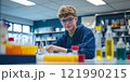 A young scientist is engaged in an exciting experiment in a colorful laboratory. The environment is bright, filled with various glassware and chemicals. Curiosity and discovery are at the heart of 121990215