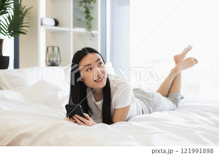 Young Asian woman using smartphone while lying on bed in cozy bright bedroom. Enjoying leisure time, communicating online. Relaxing and comfortable home environment with natural light. 121991898