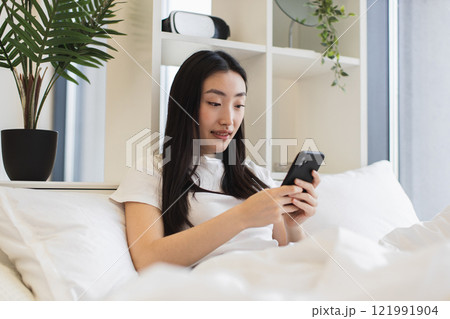 Asian woman relaxing on bed using smartphone for social media or messaging. Bright and cozy modern bedroom setting, conveying comfort and relaxation. 121991904