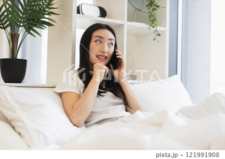 Relaxed Asian woman on bed having phone call. Young adult enjoying conversation in cozy home setting. Bright room enhances calm atmosphere. Relaxed Asian woman on bed having phone call. Young adult enjoying conversation in cozy home setting. Bright room enhances calm atmosphere. 121991908