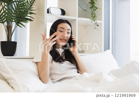 Asian woman in casual white shirt having phone call while relaxing on comfortable bed in bright room. 121991909