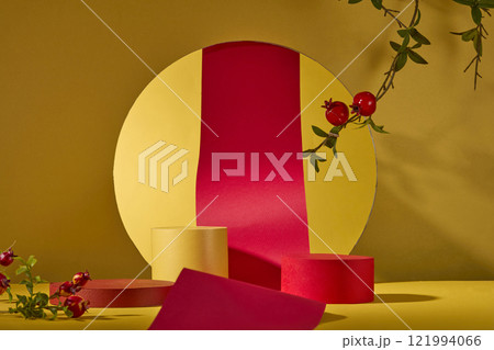 On a yellow background, a red paper roll is seen in front through a circle-shaped cutout. On a gold surface are two empty podiums for showcasing products. Some pomegranate branches as decorations. 121994066