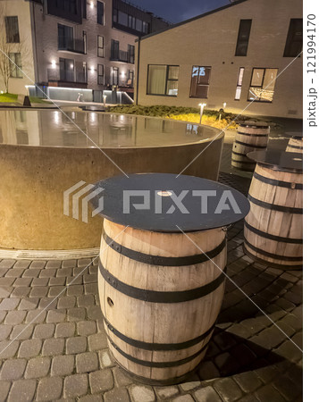 Wooden barrel tables with black tops sit beside a circular water feature at dusk. High quality photo 121994170