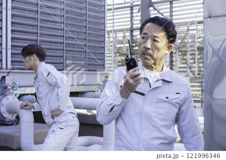 屋上の給排水設備の前でトランシーバーで話す作業服の男性 撮影協力:学校法人電波学園 東京電子專門学校 屋上の給排水設備の前でトランシーバーで話す作業服の男性 撮影協力:学校法人電波学園 東京電子專門学校 121996346
