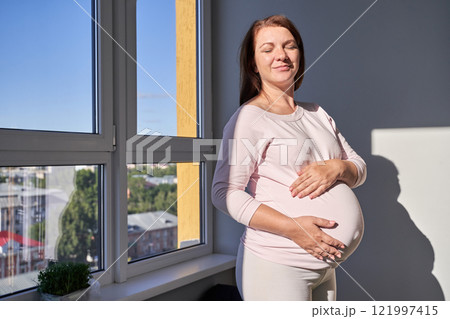 Pregnant caucasian woman relaxing by window in sunlit room embracing pregnancy 121997415